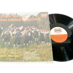 May include: A vintage vinyl record album with the title "Grüße aus dem schönen Egerland" in German. The album cover shows a band in traditional attire. The record is black, with an orange and white label. The album is from the Europa label.