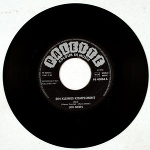 Peut inclure: Un disque vinyle noir avec du texte blanc. L'étiquette du disque indique "EIN KLEINES KOMPLIMENT" de Lou Neefs. Le disque est un single 45 tours, publié en 1961. L'étiquette présente également le logo "Palette Colour in Music".