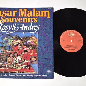 May include: A vintage vinyl record album titled "Pasar Malam Souvenirs" by Rosy & Andres. The album cover features a colorful illustration of a restaurant scene. The record itself is black with an orange label.
