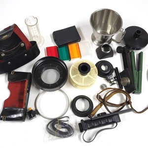 Könnte beinhalten: Eine Sammlung von Vintage-Fotozubehör für die Dunkelkammer, darunter eine schwarze Lederkameratasche, ein Film-Entwicklungsbehälter, eine Filmrolle, ein Belichtungsmesser, ein Blitzgerät und eine Vielzahl weiterer Accessoires.