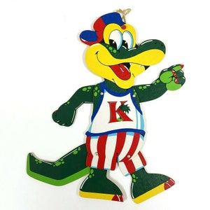 May include: A green, yellow, red and blue wooden puppet of a cartoon alligator wearing a baseball cap and a white, red and blue striped shirt with a letter K and a palm tree on it.