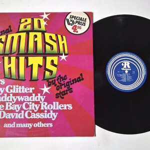 May include: A vintage vinyl record album titled "20 Smash Hits" with a pink and red cover. The album features artists like The Drifters and Gary Glitter. The black record is visible next to the album cover.