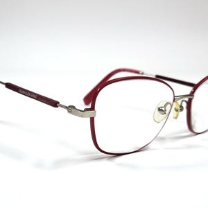 May include: Calvin Klein Jeans spectacles with a burgundy frame and silver detailing. The oval lenses are clear, and the temples display the brand name. The glasses are set against a white background.