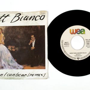 Peut inclure: Un disque vinyle vintage et sa pochette pour "More Than I Can Bear (Remix)" de Matt Bianco. La pochette présente une peinture d'un couple s'embrassant, avec un musicien jouant de la guitare. Le disque est noir.