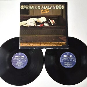 May include: Album cover for "Ópera do Malandro" by Chico Buarque, featuring a man in a white suit and hat. Two black vinyl records with blue and white labels are in the foreground. The album cover has text in Portuguese.