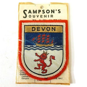 May include: A fabric badge featuring the word DEVON, a red ship on a blue background, and a red lion on a white background. The badge is mounted on a cream-colored card labeled SAMPSON'S SOUVENIR. The shield-shaped patch has a thick red border.
