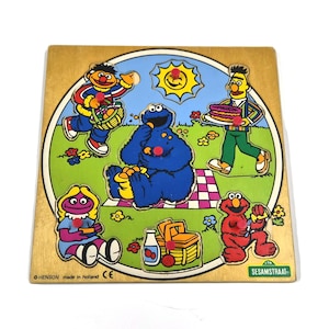 Op de afbeelding: Een houten Sesame Street puzzel met knoppen, met Ernie, Bert, Koekiemonster, Elmo en Prairie Dawn in een park. De puzzel bevat een zon, een picknickmand en een fles melk. De tekst SESAMSTRAAT staat in de rechterbenedenhoek van het bord.