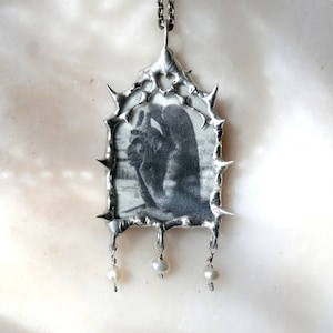 May include: A silver pendant necklace with a thorny frame and a black and white photo inside. The pendant has three pearl drops hanging from it.