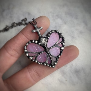 May include: A silver heart-shaped pendant necklace with a cross at the top and a pink stained glass butterfly design.