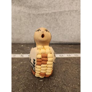 Peut inclure: Petite figurine en céramique faite à la main, avec une tête beige et une bouche ouverte. Le corps est crème avec des accents marron et beige, et des motifs géométriques noirs. La figurine mesure environ 10 cm de haut.