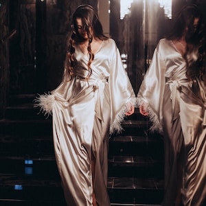 May include: A woman in a white satin robe with feather trim stands on a staircase in front of a mirror. The robe has a long, flowing silhouette and a sash tied at the waist.