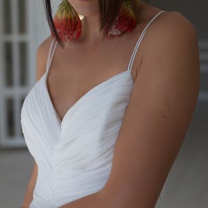 May include: A woman wearing a white, sleeveless, V-neck dress with a pleated bodice. She is wearing large, red and green floral earrings.