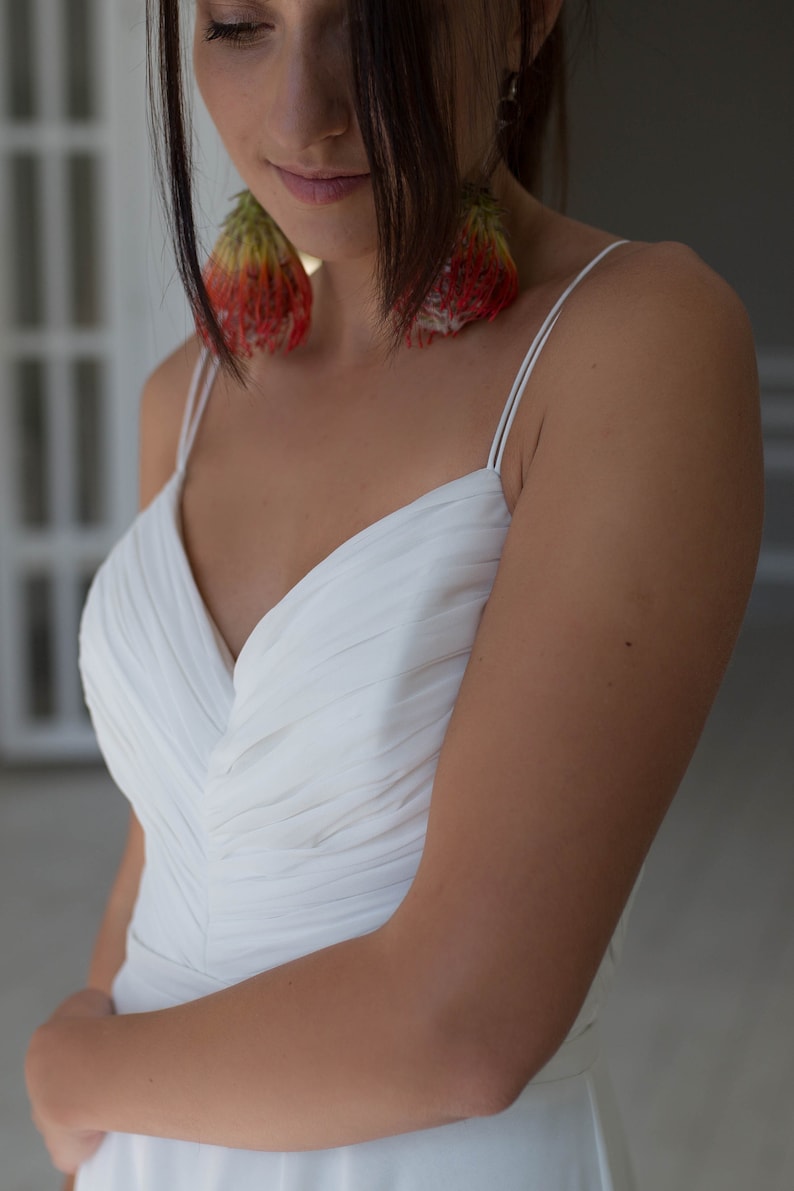 May include: A woman wearing a white, sleeveless, V-neck dress with a pleated bodice. She is wearing large, red and green floral earrings.