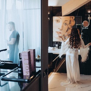 May include: A woman in a white satin robe with feather trim stands in a bathroom with a large painting of a woman's face on the wall. She is holding a glass of champagne. A man is visible through a window in the background.