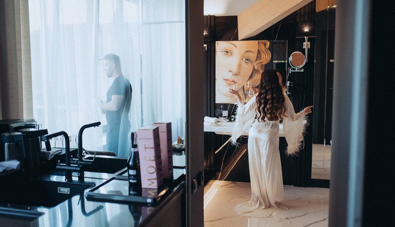 May include: A woman in a white satin robe with feather trim stands in a bathroom with a large painting of a woman's face on the wall. She is holding a glass of champagne. A man is visible through a window in the background.