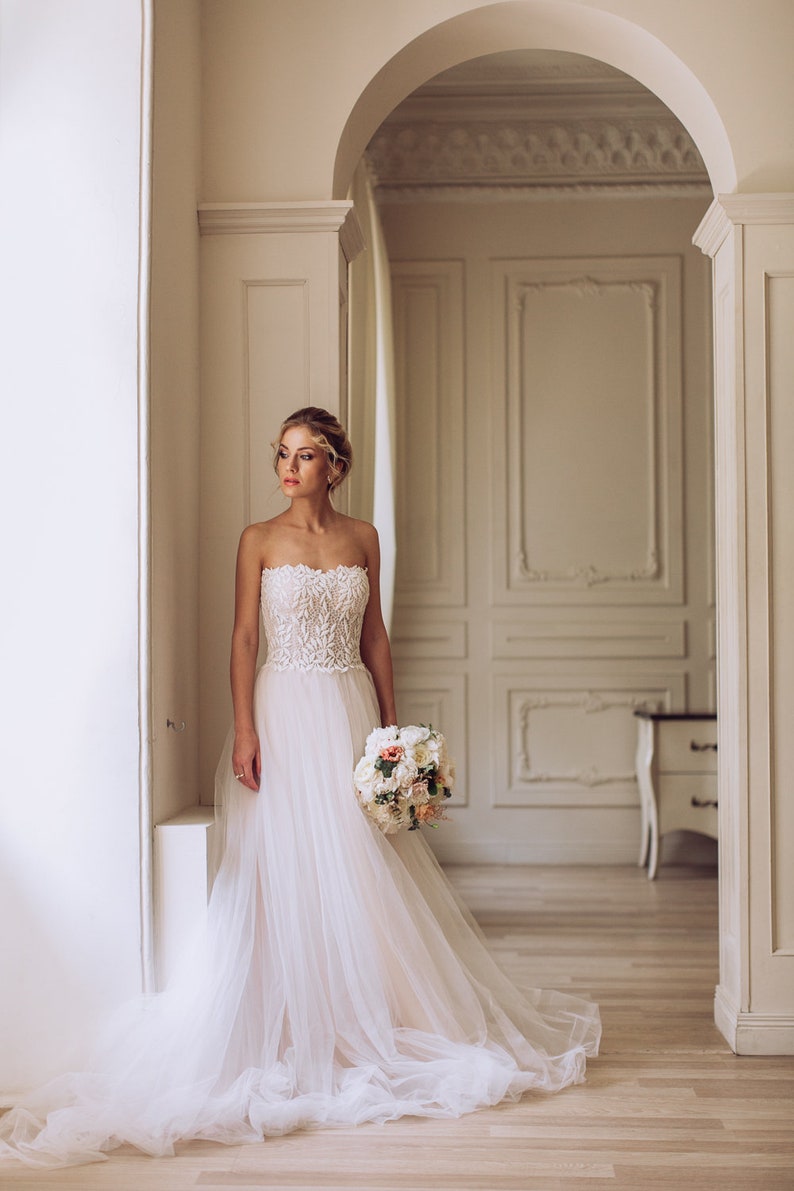 May include: A bride in a white strapless wedding dress with a tulle skirt and a bouquet of white and pink flowers. She is standing in a white room with ornate moldings.
