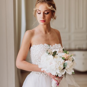 May include: A bride in a white strapless wedding dress with lace detailing holds a bouquet of white peonies and greenery.