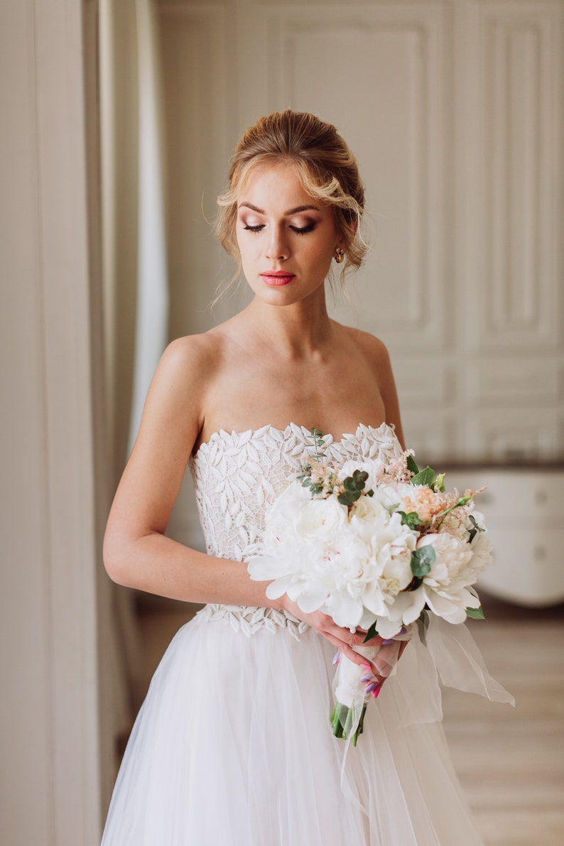 May include: A bride in a white strapless wedding dress with lace detailing holds a bouquet of white peonies and greenery.