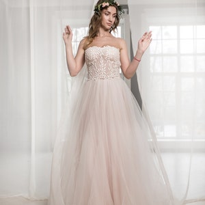 May include: A bride wearing a white strapless wedding dress with a floral crown. The dress has a lace bodice and a tulle skirt. The bride is standing in front of a white curtain.