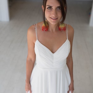 May include: A woman wearing a white, sleeveless, v-neck dress with a flowing skirt. She is wearing large, red and yellow feather earrings.