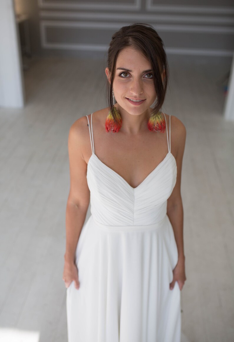 May include: A woman wearing a white, sleeveless, v-neck dress with a flowing skirt. She is wearing large, red and yellow feather earrings.