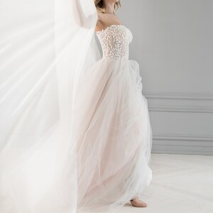 May include: A bride wearing a white strapless wedding dress with a floral lace bodice and a flowing tulle skirt. She is standing in front of a white sheer curtain and has a floral crown in her hair.