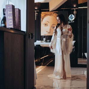 May include: A woman in a white satin robe with feather trim stands in a bathroom with a glass of red wine. She is looking down at the glass. The bathroom has a black and white marble floor and a black vanity with a white sink.
