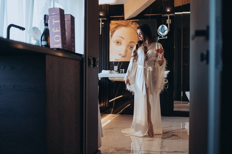 May include: A woman in a white satin robe with feather trim stands in a bathroom with a glass of red wine. She is looking down at the glass. The bathroom has a black and white marble floor and a black vanity with a white sink.