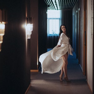 May include: A woman in a white robe walks down a hallway with dark walls and a light fixture on each side. The robe is flowing behind her as she walks.