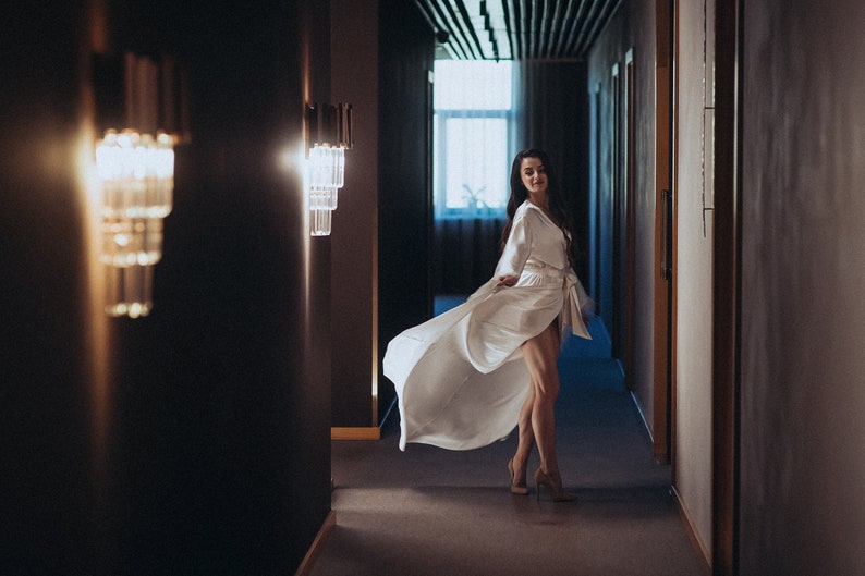 May include: A woman in a white robe walks down a hallway with dark walls and a light fixture on each side. The robe is flowing behind her as she walks.