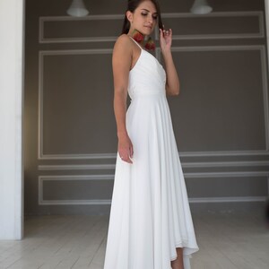 May include: A white high-low wedding dress with a simple, elegant design. The dress has a fitted bodice and a flowing skirt that is shorter in the front and longer in the back. The model is wearing red high heels.