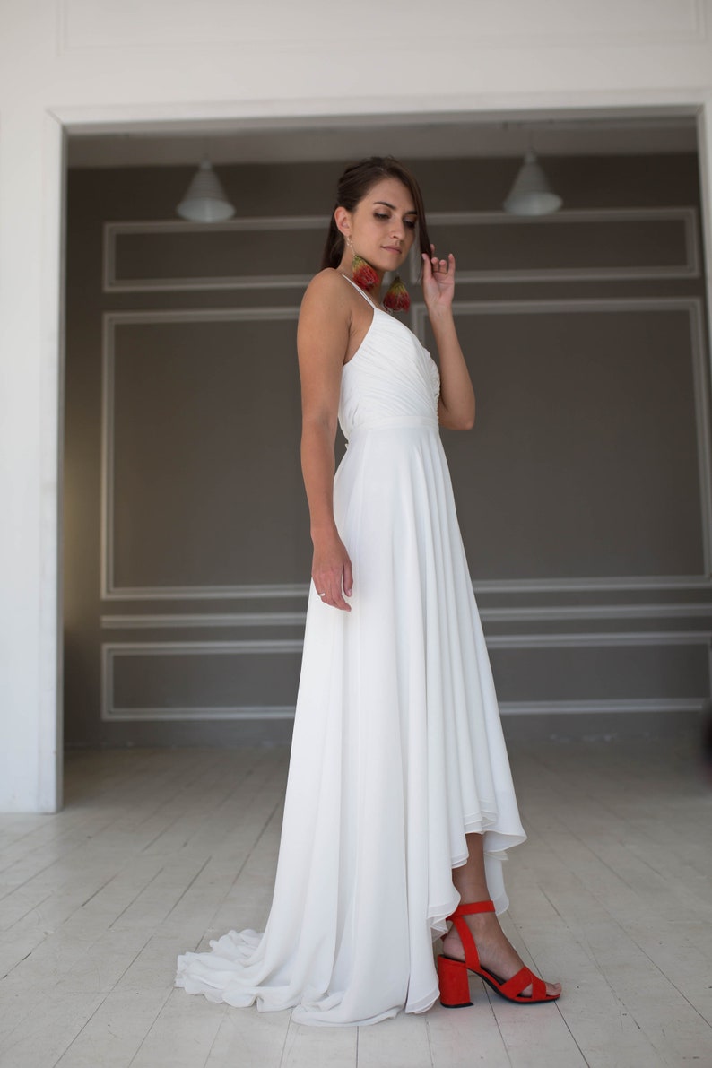 May include: A white high-low wedding dress with a simple, elegant design. The dress has a fitted bodice and a flowing skirt that is shorter in the front and longer in the back. The model is wearing red high heels.