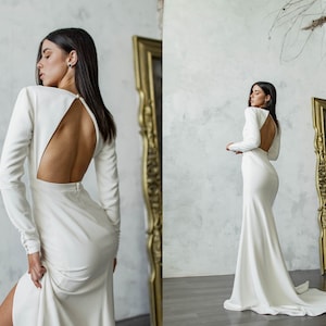 May include: A white long-sleeved wedding dress with a low back and a fitted silhouette. The dress has a slit in the front and a train.