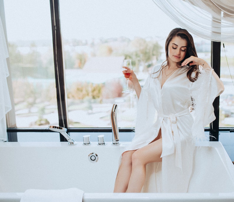 May include: A woman in a white satin robe with lace trim sits in a white bathtub, holding a glass of wine. She is looking down at the glass, and the window behind her shows a view of a city.