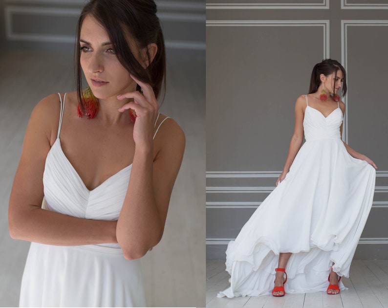 May include: A white bridal gown with a high-low hemline and spaghetti straps. The gown is made of a flowing fabric and has a simple, elegant design. The model is wearing red heels.