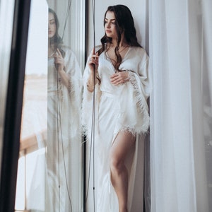 May include: A woman in a white satin robe with feather trim stands by a window. The robe has a long, flowing silhouette and a deep V-neckline.