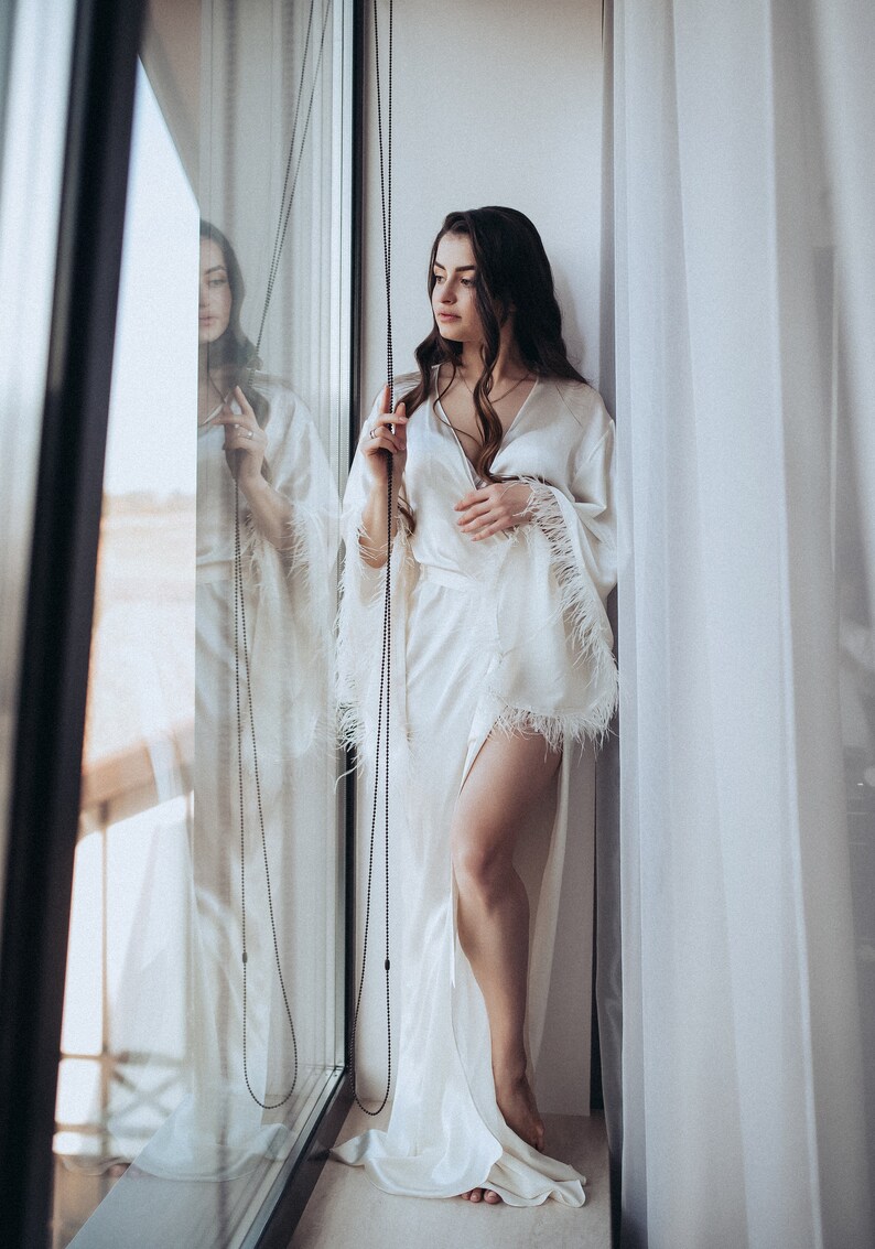 May include: A woman in a white satin robe with feather trim stands by a window. The robe has a long, flowing silhouette and a deep V-neckline.