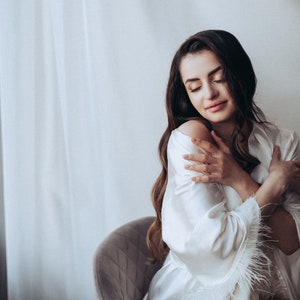 May include: A woman in a white satin robe with feather trim, sitting in a chair and holding herself. The robe has a relaxed fit and a shawl collar.