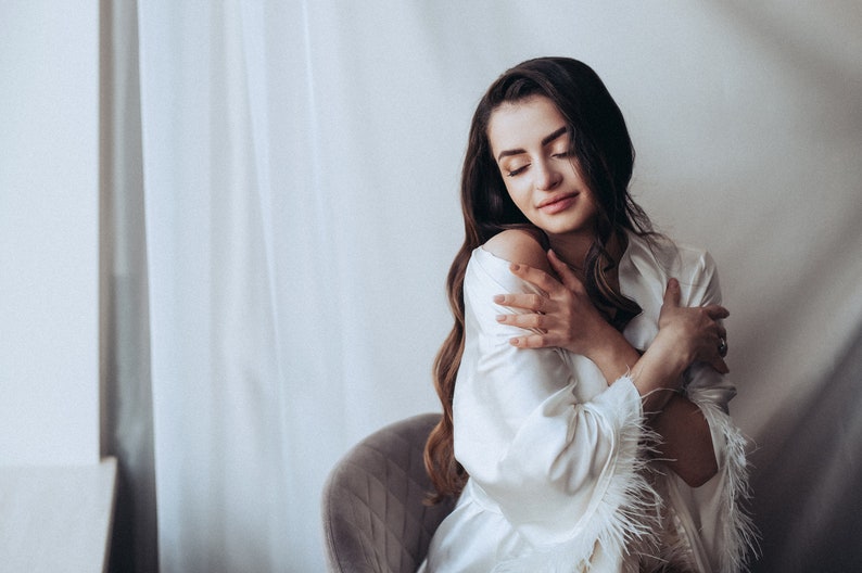 May include: A woman in a white satin robe with feather trim, sitting in a chair and holding herself. The robe has a relaxed fit and a shawl collar.