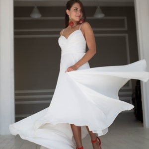 May include: A woman in a white wedding dress with a flowing skirt, standing in a white room with a doorway in the background. She is wearing red shoes and has red earrings.