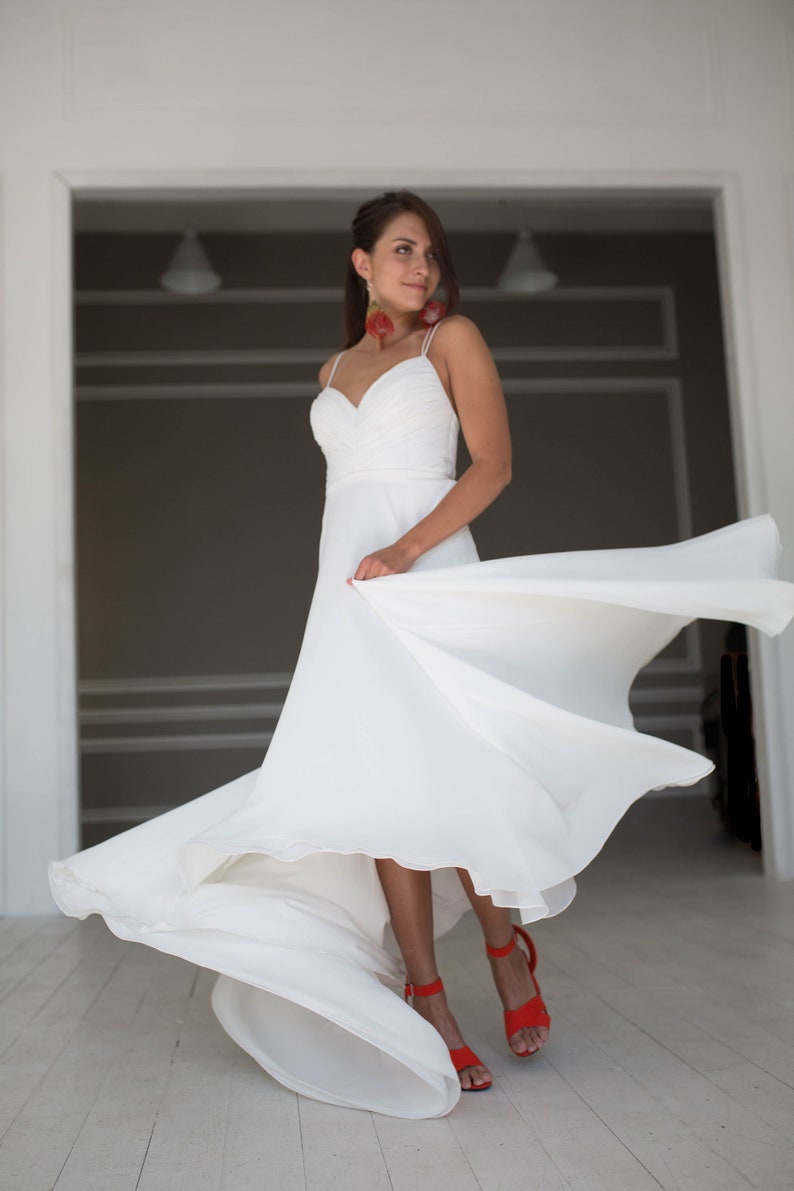 May include: A woman in a white wedding dress with a flowing skirt, standing in a white room with a doorway in the background. She is wearing red shoes and has red earrings.
