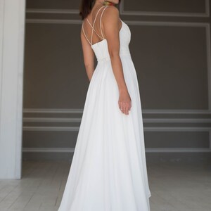 May include: A white wedding dress with a simple, elegant design. The dress has a fitted bodice with a low back and a flowing skirt.