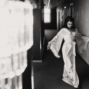 May include: A woman in a white satin robe with feather trim walks down a hallway. The robe has a long, flowing silhouette and a sash tied at the waist.