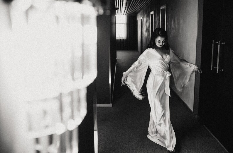 May include: A woman in a white satin robe with feather trim walks down a hallway. The robe has a long, flowing silhouette and a sash tied at the waist.