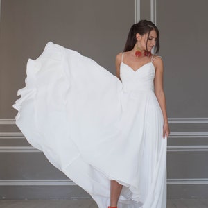 May include: A woman wearing a white wedding dress with a flowing skirt and red shoes. She is standing in a room with gray walls and white trim.