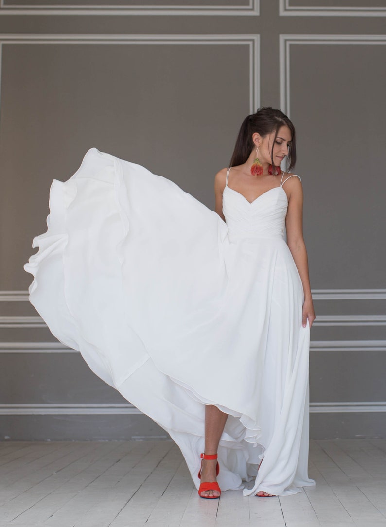 May include: A woman wearing a white wedding dress with a flowing skirt and red shoes. She is standing in a room with gray walls and white trim.