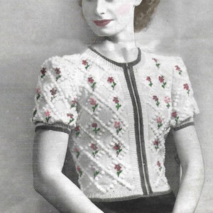 May include: A white knitted cardigan with a zipper closure and floral embroidery. The cardigan has short sleeves and a round neckline.