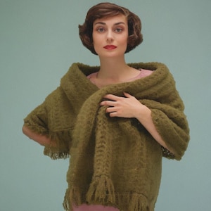 May include: An olive green shawl with fringe detail. The shawl is draped over the shoulders. The shawl is made of a textured material. The model is wearing a pink top and has short brown hair.