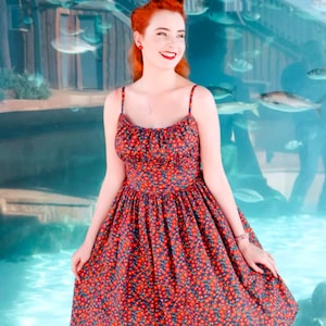 May include: A red floral print sundress with thin straps. The dress features a fitted bodice and a full skirt. The fabric has a pattern of small red flowers on a navy blue background. The dress is a vintage style.
