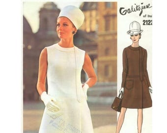 Può includere: Una busta per cartamodello Vogue Couturier Design con un abito bianco senza maniche, un cappello pillbox abbinato e guanti bianchi. L'illustrazione mostra un abito marrone a maniche lunghe con tasca, cappello coordinato e una borsa.